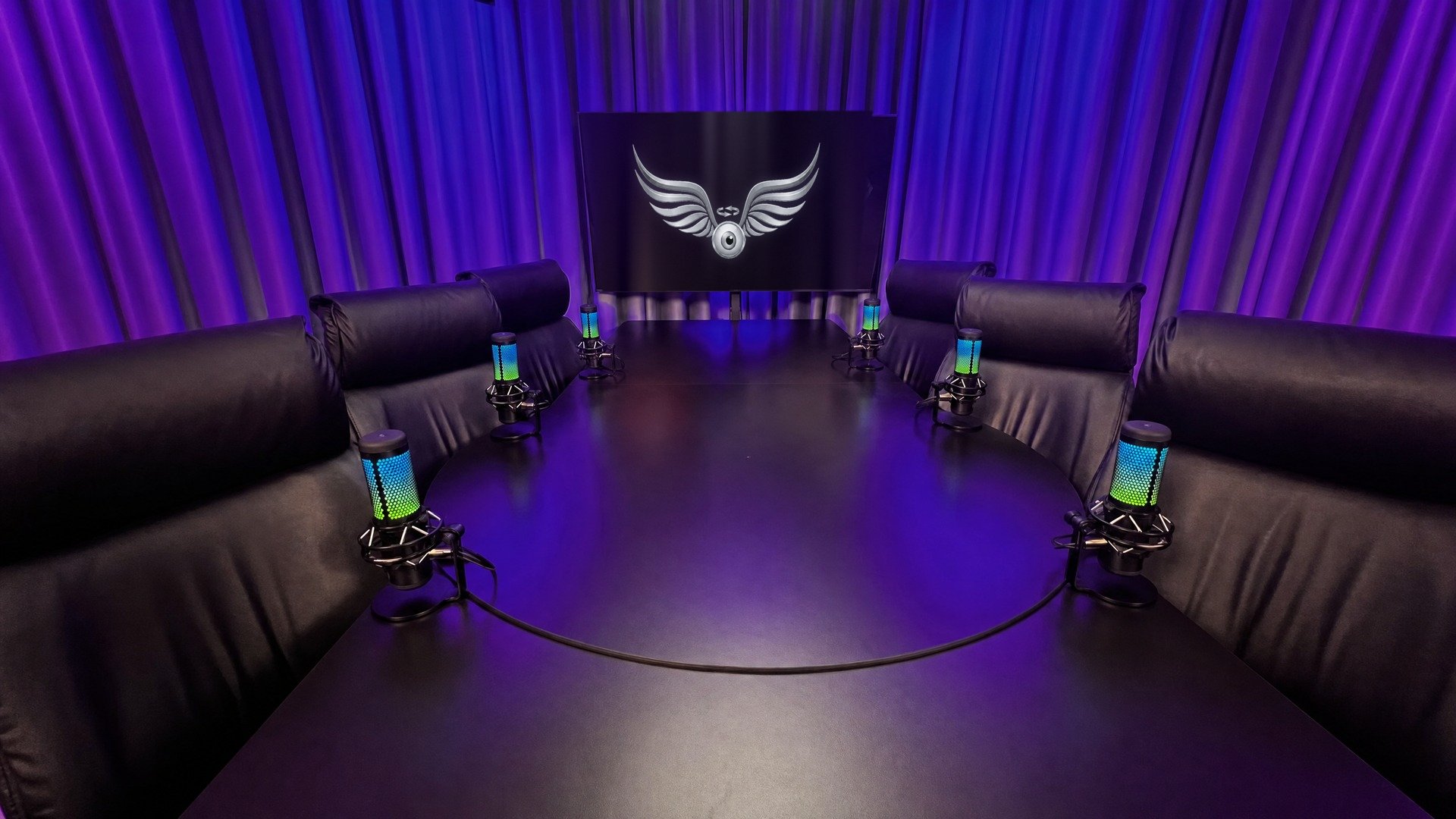 Estúdio de podcast vazio, com seis microfones ao redor de uma mesa oval e iluminação azul e violeta